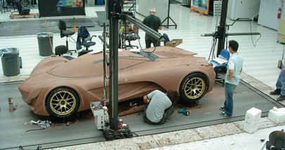 Full-scale clay car model on a surface plate with modelers working around it in a professional automotive design studio